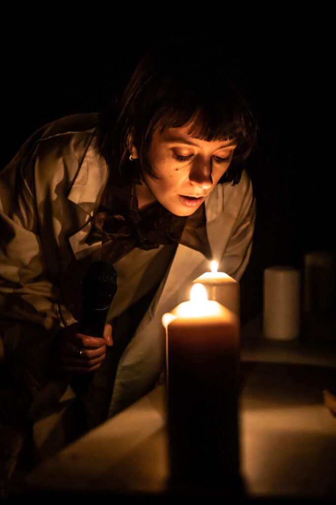 Brunette white woman in a lab coat blows out a lit candle. Darkness surrounds her.